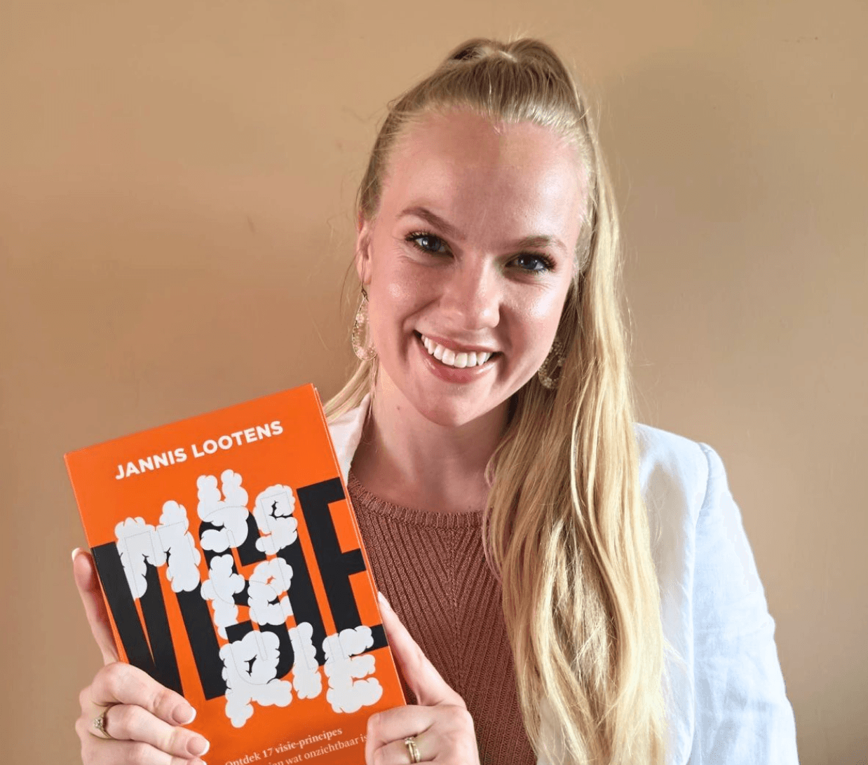 Danielle Joy Visie Mysterie Smiling woman with long blonde hair in a light blazer holding an orange book titled JANNIS LOOTENS with white abstract shapes on the cover Danielle Joy Sawicka Page 6