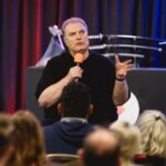 Male speaker on stage delivering a talk to an audience holding an orange microphone and gesturing with his other hand red and blue curtains behind him Jim Stovall