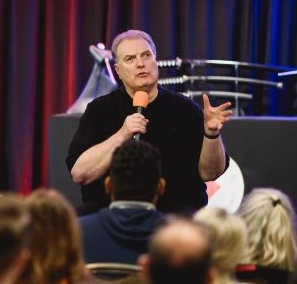 Jim Stovall Visie Mysterie Male speaker on stage delivering a talk to an audience holding an orange microphone and gesturing with his other hand red and blue curtains behind him Jim Stovall